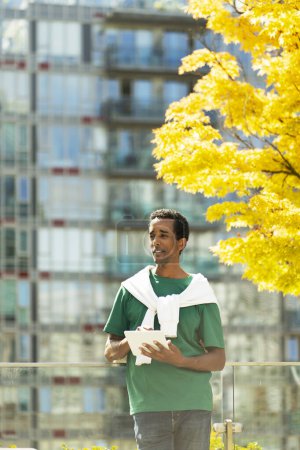 Foto de Retrato del hombre afroamericano guapo sosteniendo la tableta digital mirando hacia otro lado, de pie en la calle urbana. Estudiante atractivo estudiando, leyendo e libro. Concepto de tecnología moderna - Imagen libre de derechos