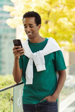 Téléchargez les photos : Portrait d'un homme afro-américain souriant attrayant tenant un téléphone portable lisant un message texte, communication en ligne dans la rue - en image libre de droit