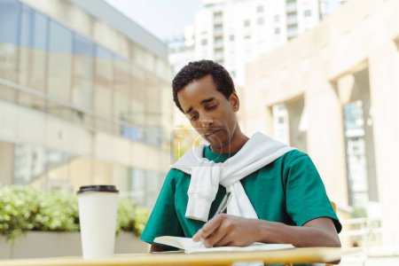 Téléchargez les photos : Portrait d'un homme afro-américain souriant, écrivain assis au bureau dans un café, pause café prenant des notes dans un journal intime à l'extérieur. Étudiant heureux intelligent étudiant assis sur le campus universitaire - en image libre de droit