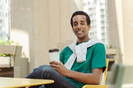 Foto de Retrato de sonriente hombre afroamericano positivo sentado en la cafetería sosteniendo taza de café con bebida, relajante, mirando a la cámara. Concepto de descanso café - Imagen libre de derechos