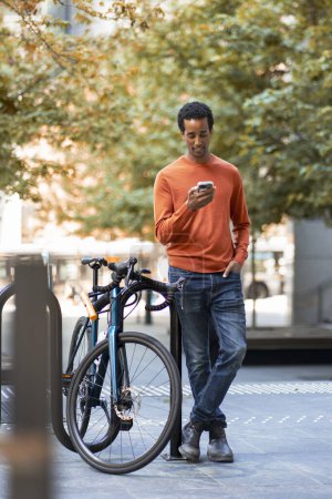 Photo for Attractive smiling African American man holding mobile phone, standing on urban street, near bicycle. Positive businessman checking mail, text message. Technology concept - Royalty Free Image