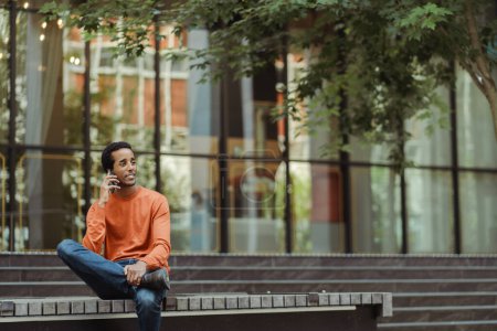 Téléchargez les photos : Portrait d'un bel homme afro-américain souriant parlant sur un téléphone portable, répondant à un appel regardant loin dans une rue urbaine, copiant de l'espace. Entreprise prospère, concept technologique - en image libre de droit
