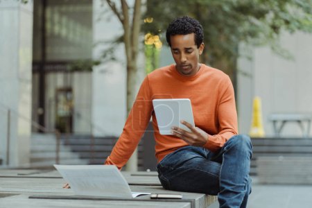 Foto de Retrato del hombre afroamericano sosteniendo una tableta digital, leyendo un libro electrónico, sentado en la calle urbana, al aire libre, espacio para copiar. Hombre de negocios guapo revisando correo, mensaje de texto. Concepto educativo - Imagen libre de derechos