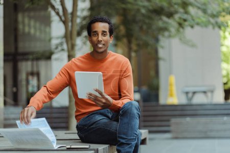 Foto de Retrato del hombre afroamericano sosteniendo una tableta digital, mirando a la cámara, sentado en la calle urbana, al aire libre, espacio para copiar. Hombre de negocios guapo revisando correo, mensaje de texto. Concepto educativo - Imagen libre de derechos