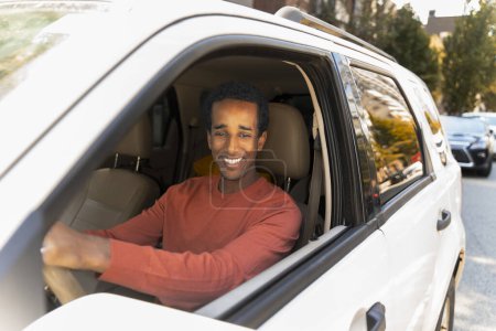 Foto de Retrato del joven conductor afroamericano sonriente con ropa casual sentado detrás del volante, conduciendo, mirando en el espejo retrovisor. Concepto de movimiento, vehículo - Imagen libre de derechos