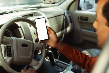 Téléchargez les photos : Jeune homme afro-américain tenant un téléphone portable avec écran blanc vierge, maquette, endroit pour le texte, assis dans la voiture. Concept technologique - en image libre de droit