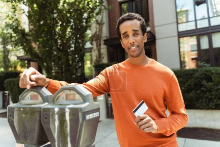 Foto de Retrato del atractivo hombre afroamericano sonriente parado en el parquímetro, sosteniendo la tarjeta de crédito, pagando por el estacionamiento, mirando a la cámara al aire libre. Concepto de transporte - Imagen libre de derechos