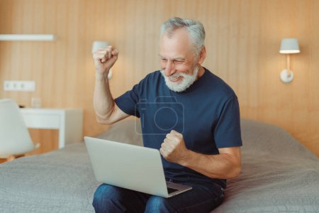Photo for Portrait of smiling senior man with beard sitting on bed using laptop in living room, making winner gesture. Attractive elderly writer working at computer at home. Remote job concept - Royalty Free Image