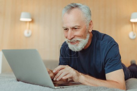 Photo for Portrait of attractive smiling gray haired senior man with beard using laptop, lying on bed in hotel. Handsome, successful elderly pensioner playing game in living room. Concept of working from home - Royalty Free Image