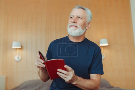 Foto de Atractivo hombre mayor positivo rima libro mientras está sentado en la cama en el hotel, mirando hacia otro lado. Guapo abuelo canoso leyendo. Concepto educativo - Imagen libre de derechos