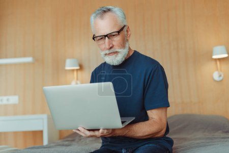Téléchargez les photos : Portrait d'un homme âgé aux cheveux gris portant des lunettes, assis sur son lit dans le salon, à l'aide d'un ordinateur portable, d'une machine à écrire, d'un travail à distance. Programmeur senior professionnel travaillant à la maison. Concept technologique - en image libre de droit