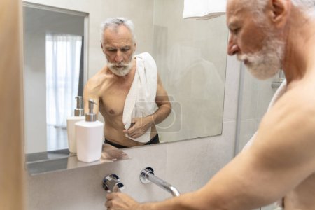 Téléchargez les photos : Portrait d'un bel homme âgé debout dans la salle de bain tenant une serviette, se lavant. Homme âgé attrayant avec torse nu, procédures d'hygiène torse nu. Concept de routine matinale - en image libre de droit