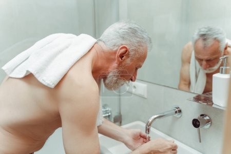 Téléchargez les photos : Portrait d'homme aux cheveux gris retraité sérieux avec serviette, torse nu se lavant dans le lavabo, eau coulant du robinet, dans la salle de bain de l'appartement. Concept de procédures hygiéniques, routine matinale - en image libre de droit