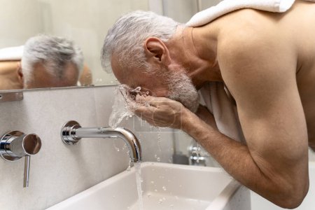 Foto de Hombre mayor de pelo gris con toalla torso desnudo lavándose la cara en el baño. Concepto de rutina matinal - Imagen libre de derechos