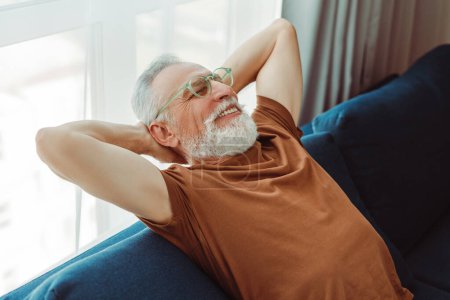 Foto de Retrato de un anciano barbudo sonriente sentado en un cómodo sofá, relajándose en casa - Imagen libre de derechos