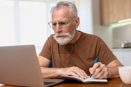 Téléchargez les photos : Portrait d'un homme âgé barbu élégant portant des lunettes, assis au bureau, utilisant un ordinateur portable, prenant des notes, enseignant en ligne, travaillant à la maison. Retraité attrayant travaillant en ligne. Concept d'emploi à distance - en image libre de droit