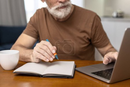 Photo for Closeup, gray haired bearded senior man working at workplace, using laptop, taking notes, selective focus on hand, holding pen. Mature freelancer remote job from home - Royalty Free Image