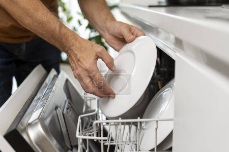 Téléchargez les photos : L'homme charge la vaisselle dans le lave-vaisselle, met les plaques, gros plan, sélectionne la mise au point. Nettoyant nettoyage cuisine dans l'appartement. Concept technologique - en image libre de droit