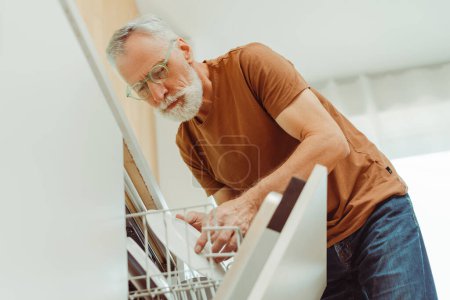 Téléchargez les photos : Portrait d'un bel homme âgé portant des lunettes et des vêtements décontractés chargeant un lave-vaisselle avec de la vaisselle dans la cuisine à la maison. Homme âgé attrayant engagé dans les tâches ménagères. Concept de travaux ménagers - en image libre de droit