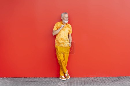 Téléchargez les photos : Beau vieil homme sérieux, hipster barbu portant des vêtements orange élégant en attente, debout dans la rue, isolé sur fond rouge. Modèle de mode posant pour des photos à l'extérieur, détournant les yeux - en image libre de droit