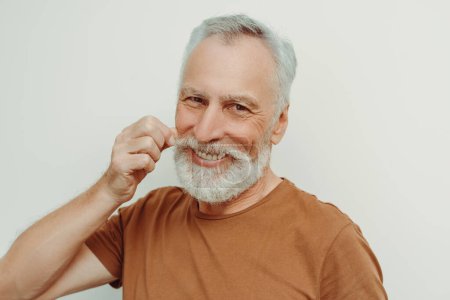 Foto de Atractivo hombre mayor sonriente con barba, fijando bigote mirando a la cámara aislada sobre fondo gris. Hombre guapo con peinado elegante en camiseta marrón posando para la imagen. Concepto de publicidad - Imagen libre de derechos