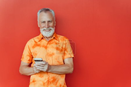 Téléchargez les photos : Homme âgé souriant tenant téléphone mobile shopping en ligne sur la rue debout près du mur rouge, copier l'espace. Joyeux beau hipster barbu en utilisant smartphone, communication en ligne. Services bancaires mobiles - en image libre de droit