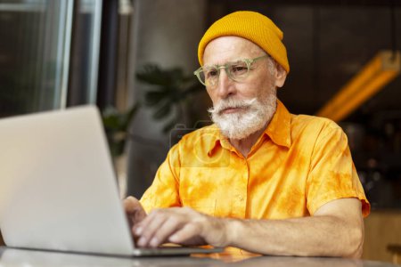 Photo for Handsome senior man, bearded programmer wearing yellow hat using laptop computer working online at workplace. Stylish freelancer, copywriter typing on keyboard sitting in modern cafe. Remote job - Royalty Free Image