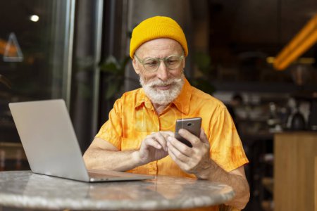 Photo for Bearded smiling senior man holding mobile phone using modern app shopping online sitting in cafe. Happy handsome freelancer receive payment, mobile banking. Technology, remote job concept - Royalty Free Image