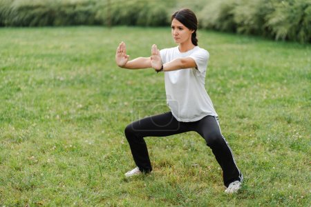 Téléchargez les photos : Femme sérieuse s'entraînant, pratiquant le wushu dans le pré vert du parc. Style de vie sain, kungfu, concept d'arts martiaux - en image libre de droit