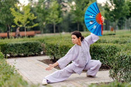 Foto de Mujer caucásica con abanico usando kimono practicando wushu en el parque. Artes marciales chinas, concepto de estilo de vida saludable - Imagen libre de derechos