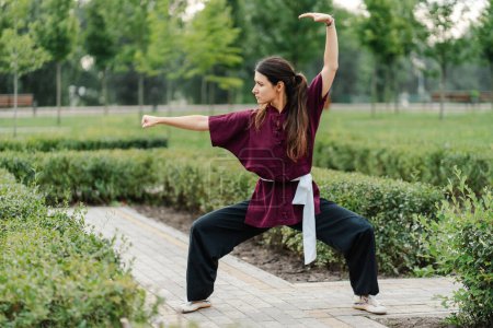 Foto de Wushu maestro en kimono uniforme de entrenamiento en el césped del parque. Campeón de Kungfu entrena las artes marciales en la naturaleza en el fondo de los árboles - Imagen libre de derechos