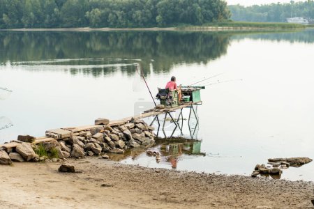 Foto de Hombre sentado silla y caña de pescar en el lago. Pescador disfrutando de pasatiempo favorito en la orilla del lago. Concepto de supervivencia - Imagen libre de derechos