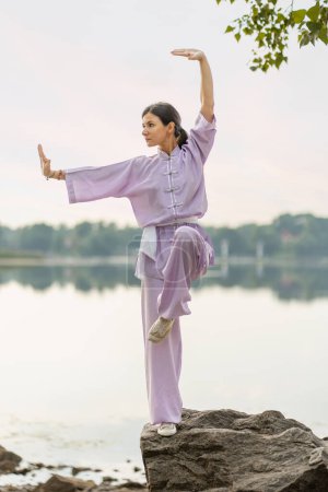 Photo for Serious woman kungfu master wearing kimono training, practicing wushu standing on stone near water. Healthy lifestyle, martial arts concept - Royalty Free Image