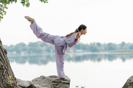Photo for Serious woman kungfu master wearing kimono training, practicing wushu standing on stone near water, copy space. Healthy lifestyle, martial arts concept - Royalty Free Image