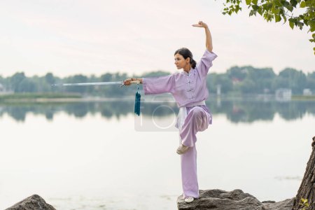 Téléchargez les photos : Maître Kungfu femme portant kimono tenant épée, pratiquant wushu debout sur la pierre près de l'eau, copier l'espace. Mode de vie sain, concept des arts martiaux - en image libre de droit