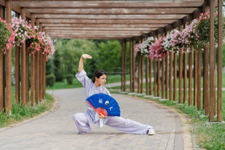 Photo for Caucasian woman with fan wearing kimono posing at at green park after practicing wushu. Chinese martial arts, healthy lifestyle concept - Royalty Free Image