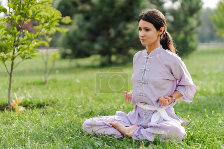 Photo for Portrait of serious woman wearing kimono sitting at the lotus pose, practicing wushu in park. Healthy lifestyle, kungfu, martial arts concept - Royalty Free Image