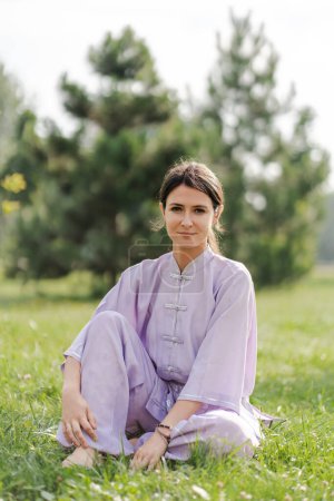 Photo for Wushu woman wearing kimono sitting at the grassy lawn and looking at camera while relaxing after training. Healthy lifestyle concept - Royalty Free Image