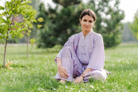 Téléchargez les photos : Femme Wushu portant kimono assis à la pelouse herbeuse et regardant la caméra tout en se relaxant après l'entraînement. Concept de mode de vie sain - en image libre de droit