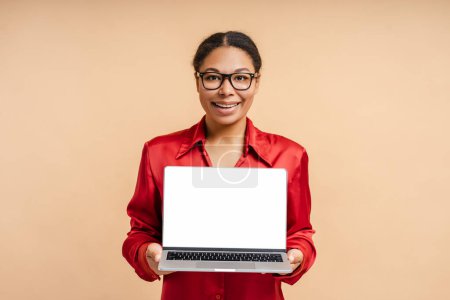 Téléchargez les photos : Portrait de femme afro-américaine souriante tenant un ordinateur portable avec écran blanc regardant la caméra isolée sur fond beige - en image libre de droit