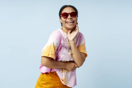 Téléchargez les photos : Portrait de belle femme afro-américaine riante portant un t-shirt teint cravate à la mode et des lunettes de soleil isolées sur fond bleu. Modèle de mode élégant souriant s'amusant posant pour l'image en studio - en image libre de droit