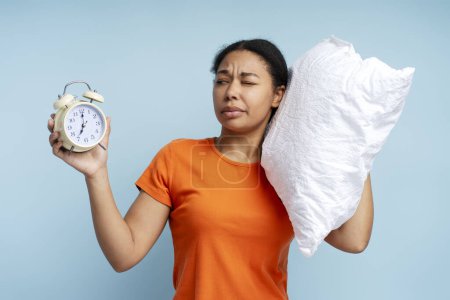 Photo for Calm young sad woman relaxing at home holding in hand pillow and alarm clock isolated on blue background studio portrait. Bad nap concept - Royalty Free Image