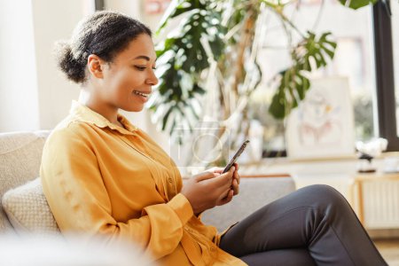 Téléchargez les photos : Femme d'affaires afro-américaine souriante moderne, pigiste tenant un téléphone mobile, travaillant en ligne depuis la maison. Élégant rédacteur avec smartphone assis dans un café. Une entreprise prospère - en image libre de droit