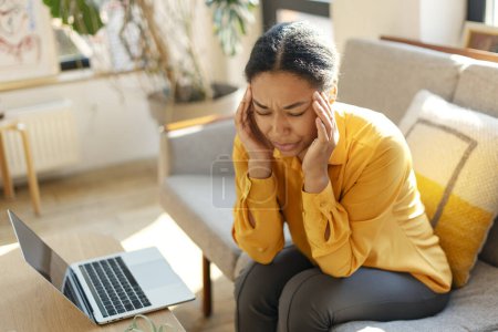 Foto de Mujer de negocios afroamericana cansada frotando templos mientras trabaja portátil en línea desde casa. Elegante dama sentada en la cafetería. Negocios exitosos - Imagen libre de derechos