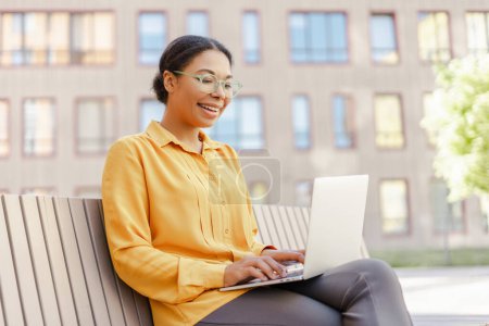 Foto de Hermosa mujer afroamericana sonriente, trabajadora sosteniendo computadora portátil, sentada en el banco al aire libre. Tecnología moderna, videoconferencia, concepto de lección en línea - Imagen libre de derechos