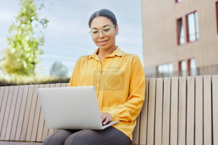 Téléchargez les photos : Belle femme afro-américaine souriante, travailleuse tenant un ordinateur portable, bénéficiant d'une éducation à distance sur un banc à l'extérieur. Technologie moderne, vidéoconférence, concept de leçon en ligne - en image libre de droit
