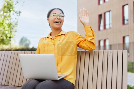 Foto de Mujer de negocios afroamericana sonriente sosteniendo una computadora portátil, saludando con la mano en el banco. Tecnología moderna, concepto de trabajo remoto - Imagen libre de derechos