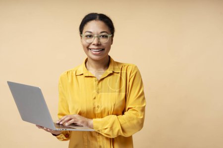 Téléchargez les photos : Belle femme d'affaires afro-américaine souriante, gestionnaire, pigiste tenant ordinateur portable travaillant en ligne, tapant sur le clavier isolé en arrière-plan. Étudiant intelligent portant des lunettes étude, éducation - en image libre de droit