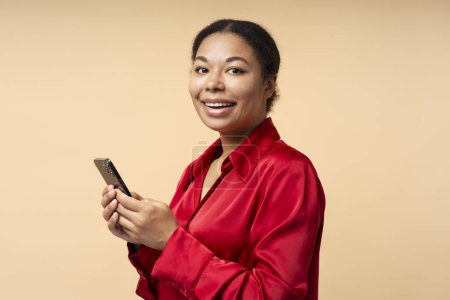 Foto de Retrato de afroamericano sonriente sosteniendo compras de teléfonos móviles en línea aislado sobre fondo beige, espacio para copiar. Empresaria exitosa usando smartphone, banca móvil - Imagen libre de derechos