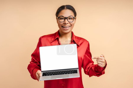 Foto de Retrato de una mujer afroamericana sonriente sosteniendo una computadora portátil apuntando con el dedo a la pantalla blanca mirando a la cámara aislada sobre un fondo beige - Imagen libre de derechos